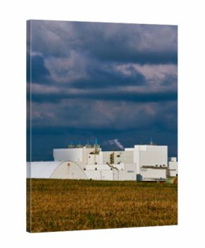 Round hay bales on farmland in Saskatchewan, Canada, with large white industrial buildings and storage domes in the background under dramatic cloudy sky.
