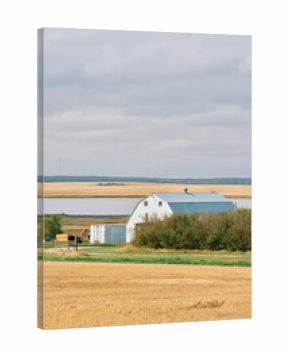 A white farm warehouse and silver grain bins stand beside harvested fields and a water body under cloudy skies in Saskatchewan, Canada.