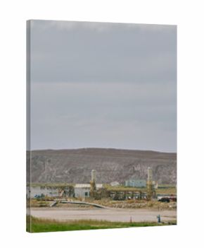 A red crane operates at an industrial facility near a potash mine in Saskatchewan, Canada, with pipelines, storage buildings, tailings pile, and a rocky embankment by the water.