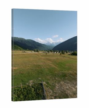A tranquil rural landscape features an expansive green field bordered by distant mountains under a bright blue sky, with a rustic fence in the foreground.