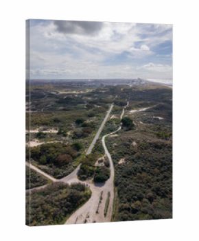 Aerial view of a coastal zone with winding paths through sandy dunes and dense vegetation, leading toward a shoreline cityscape beside the ocean under a partly cloudy sky.