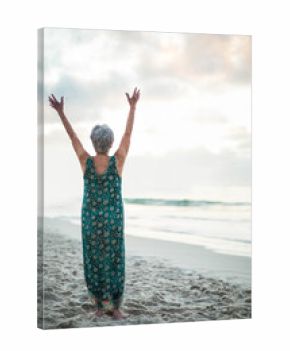 Barefoot senior woman raising arms toward sky on sandy beach by calm sea, under cloudy sky