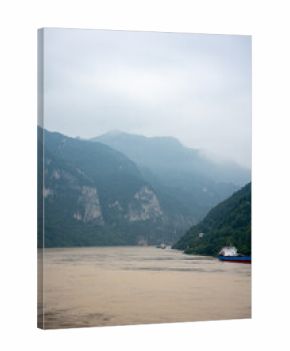Yichang Three Gorges in China features a wide river flowing between immense, green mountains under a cloudy sky with boats navigating the water.