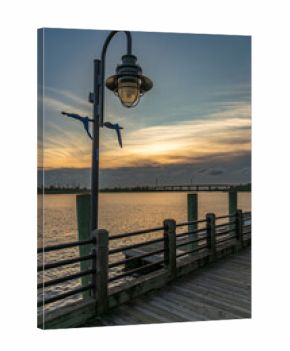A peaceful sunset illuminates the Wilmington, North Carolina Riverwalk, where a vintage lamppost stands over a wooden boardwalk beside the calm Cape Fear River.