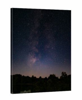 Stunning night landscape with the Milky Way stretching across the starry sky above a calm lake and dark forest silhouettes