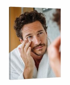young man taking care of his undereye wrinkles putting anti aging eye moisturizer handsome guy applying moisturizer and looking at himself while standing in front of the mirror in the bathroom  no lo
