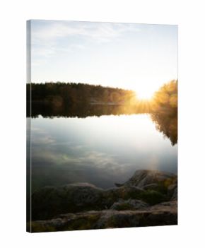 Idyllic landcsape in sweden with a calm lake reflecting the surrounding forest , a rocky foreground, golden light illuminating the scene