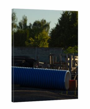 blue pipe stacked in industrial yard under warm dusk light, corrugated surface catching soft highlights, scattered pallets and fencing framing background, tall trees and concrete wall