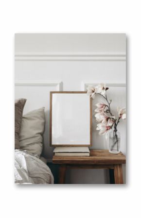 Blank wooden picture frame mockup on old book. Wooden night stand with fluted glass vase. Blooming magnolia tree branches. Scandinavian interior. Elegant bedroom. White wall background, stucco decor.