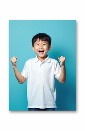 Joyful asian little boy wearing white t-shirt celebrates victory. Korean child with smile in excitement against blue background. Happiness fun. Studio portrait.