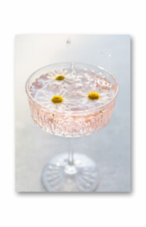 Elegant crystal glass filled with refreshing pink beverage garnished with floating daisies. Droplets splashing in the glass, creating a fresh, summery atmosphere against a soft light background.