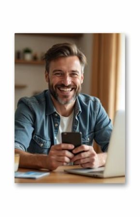 Smiling mid-adult man uses mobile phone, laptop at home dining table. Holds credit card, suggesting online shopping financial transactions. Casual domestic scene shows happy lifestyle with technology.