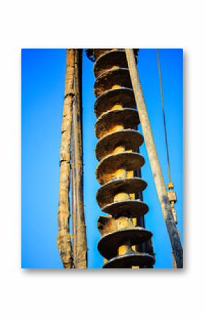 Vertical view of a large industrial auger drill used in construction and earth drilling. The image captures the full length of the spiral drill bit with surrounding cables