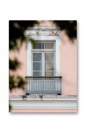 Ornate balcony with a vintage window on a pastel pink building facade, framed by blurred greenery