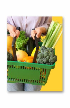 Woman holding green shopping basket with different fresh products on yellow background