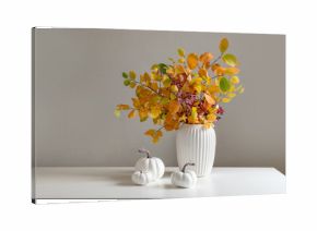 Bouquet of dry colorful red and orange fall leaves and branches in white vase and pumpkins on white table. Minimal modern composition. Autumn season. Home interior and season decoration.