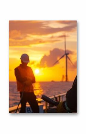 Silhouetted workers on a ship deck oversee sunset-lit wind turbines, embodying a blend of technology and nature's beauty over the ocean.