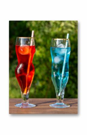 Two artistic glasses shaped like a nude woman and man filled with red and blue alcoholic cocktails on table with lush green garden background, closeup. Playful summer vibe for creative drink concepts