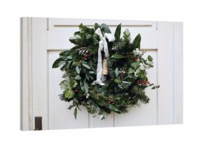 Beautiful Christmas wreath with greenery, eucalyptus, holly and red berries hanging on white door. Rustic ribbon, vintage bell. Natural festive floral decoration for winter holiday season, advent.