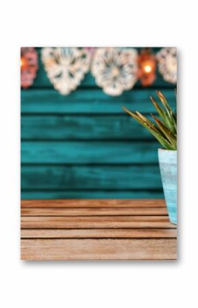 Aloe plant in a rustic turquoise pot on a wooden table with decorative papel picado and lights in the background. Mexican decor and celebration concept