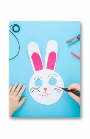 Little child hand drawing rabbit paper mask with marker on light blue table background. Pastel color. Point of view shot. Closeup. Creating easter decoration. Top down view.