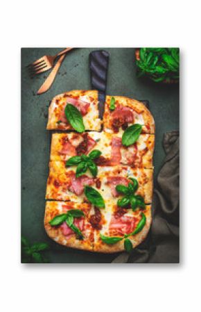 Hot Roman pizza with ham, pancetta, mozzarella cheese, sun dried tomatoes, sauce and green basil, rustic green table background, top view