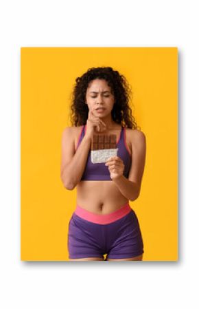 Thoughtful young African-American woman with sweet chocolate bar on yellow background