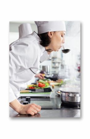 In a busy restaurant kitchen, chef tastes a steaming pan of soup, ensuring perfect seasoning of a great gourmet dish. Culinary workspace stocked with kitchen tools and fresh ingredients.