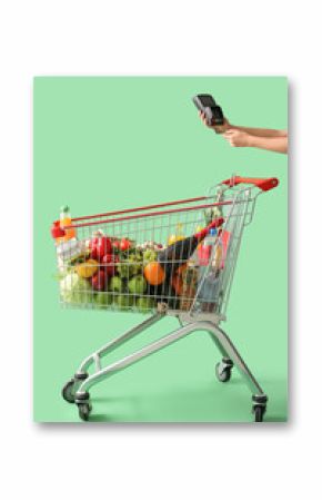 Female hand with payment terminal near shopping cart full of food on green background
