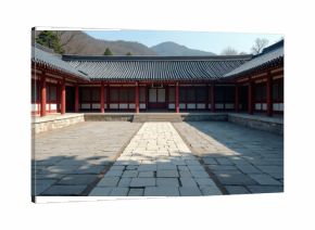 Traditional Korean house courtyard features stone pathways and tiled roofs. Red columns support covered walkways around a central stone patio. Mountains rise in the background under a clear blue sky.