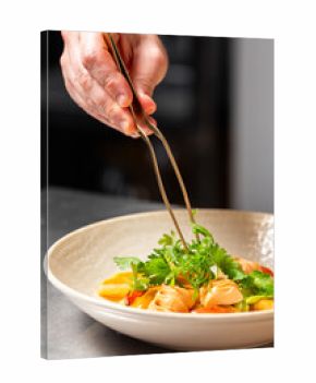 Chef plating pasta with herbs using precision tongs in a professional kitchen. Gourmet dish with creamy sauce, vegetables, and fine dining presentation