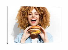 Joyful woman with curly hair holding a delicious burger, exuding happiness and enthusiasm against a plain background