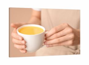 Young woman holding cup of tasty turmeric drink on beige background, closeup