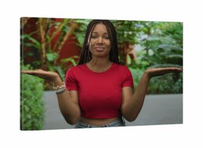 Young woman palms up presenting in a forest park area with green plants and casual red top and jeans  contemplation choice balance.