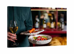 A person holds two plates of fine dining food and a glass of white wine in a restaurant setting. Focus is on the food and hands