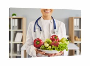 Nutritionist doctor holds vegetables promoting healthy diet. Smiling clinician with stethoscope offers a bowl of fresh salad in medical clinic. Promotes wellness healthcare and nutrition