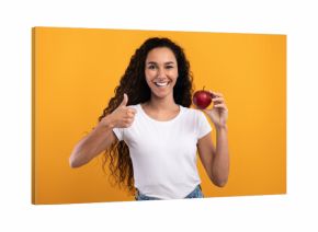 A happy millennial woman with curly hair stands against a yellow-orange background. She holds a red apple in one hand and gives a thumbs up with the other. She promotes healthy eating.