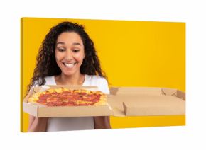 A joyful young lady gazes at an open pizza box filled with tasty Italian pizza. She stands in front of a bright yellow-orange wall, celebrating her food delivery.
