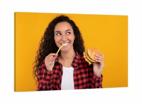 A joyful woman bites into a delicious burger, holding a fry in her other hand. She looks excited, smiling against a vibrant yellow-orange background, ready to savor fast food.