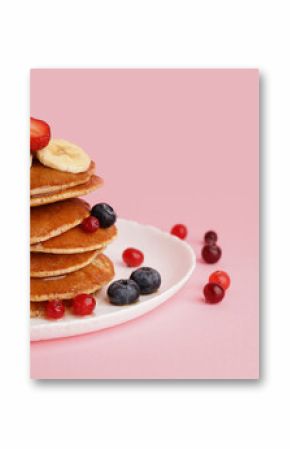 Plate with tasty pancakes, banana slices, cranberries and blueberries on pink background