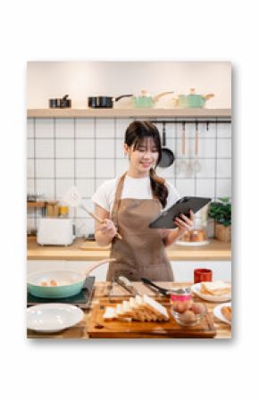 A beautiful Asian woman is looking at a recipe on her digital tablet while cooking in the kitchen.