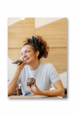 Portrait of young woman sitting in fast food restaurant with doughnut in hands and sending voice messages.