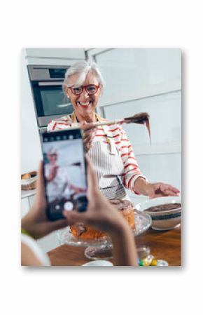 Cute granddaughter taking photos with her smartphone while grandmother making tasty chocolate cake in the kitchen.
