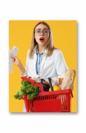 Shocked young woman with shopping basket of food and receipts on yellow background. Price rise concept