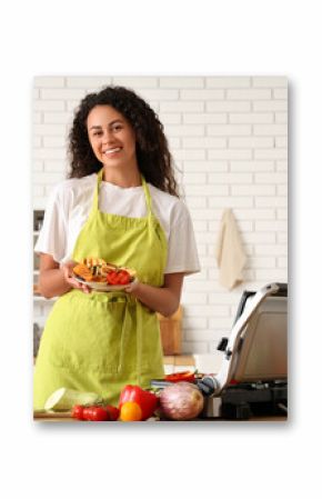 Beautiful young happy African-American woman holding plate with tasty vegetables near modern electric grill in kitchen