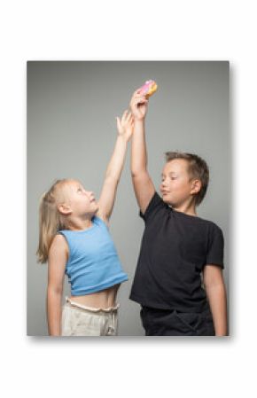 Playful sister and brother having fun holding donuts standing against white wall. Child and chocolate glazed donut