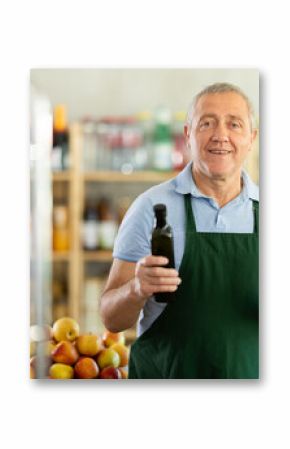 Elderly male seller at counter offering olive oil in bottle in grocery store