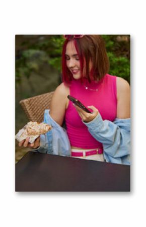 Young woman taking a picture with smartphone of a croissant at an outdoor cafe. Concept of social food culture, Gen Z dining, influencer marketing, casual lifestyle branding.