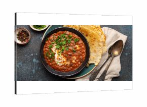 Traditional Indian Punjabi dish Dal makhani with lentils and beans in black bowl served with naan flat bread, fresh cilantro and two spoons on blue concrete rustic table top view.