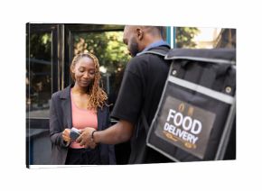 Black client completes a fast food purchase with contactless payment at the doorstep, courier holds pos terminal steady as the electronic transaction is processed. Wireless purchase.